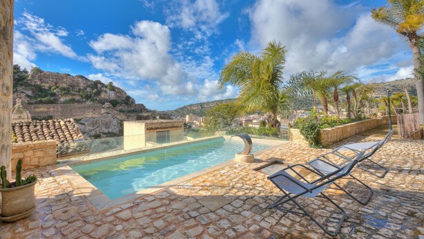 Uma piscina com um terraço de pedra, espreguiçadeiras e montanhas ao fundo.