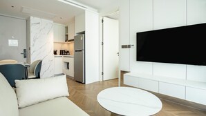 A modern living room with a sofa, a round table, a flat-screen TV mounted on the wall, and a kitchen area in the background. - Unit 2303 Beachfront | Pool-Facing + Sea View (Choeng Thale)