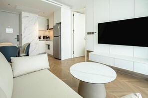 A modern living room with a sofa, a round table, a flat-screen TV mounted on the wall, and a kitchen area in the background. - Unit 2303 Beachfront | Pool-Facing + Sea View (Choeng Thale)