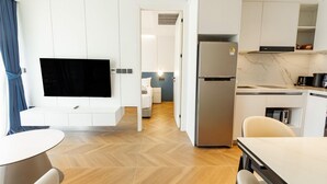 Ett modernt hotellrum med en platt-TV, en matplats med bord och stolar, ett kök med kylskåp och en hall som leder till sovrummet.