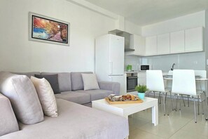 A modern living room with a grey sofa, a coffee table with muffins, and a kitchen in the background. - Panorama View Apartment In Saranda - 271, Sarandë, Albania (Sarandë)
