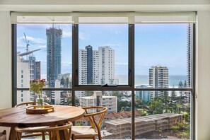 A room with a large window, a wooden table with a vase, and a view of high-rise buildings and a beach. - Skyline & Ocean View Retreat - GC 1303 (Surfers Paradise)