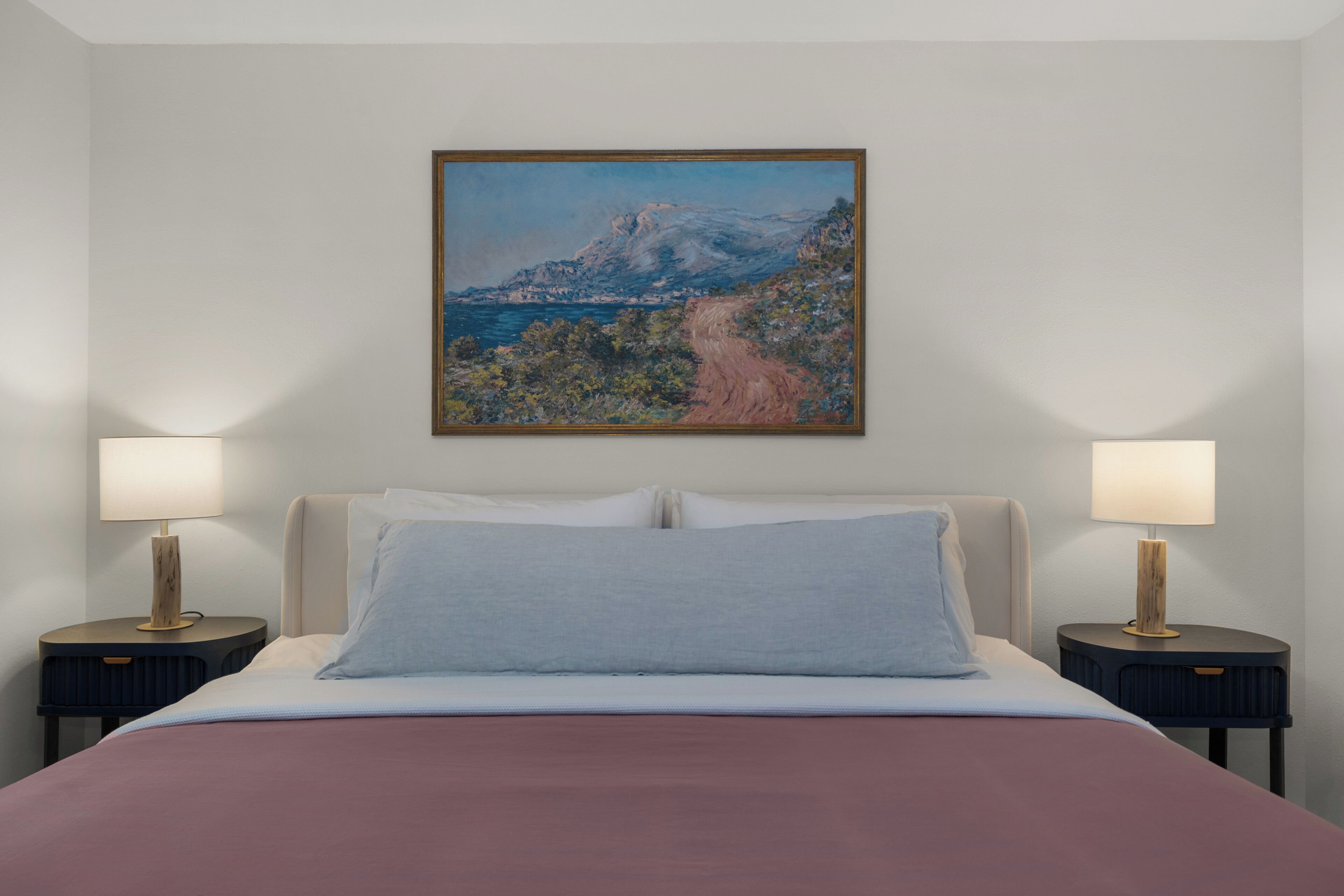 A bedroom with a bed, two bedside tables with lamps, a framed landscape painting on the wall, and a view of a mountain and sea.