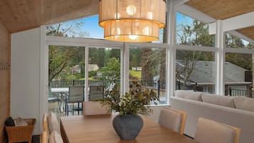 Un coin repas avec une table en bois, des chaises, un vase de fleurs et une grande fenêtre donnant sur une maison et des arbres.