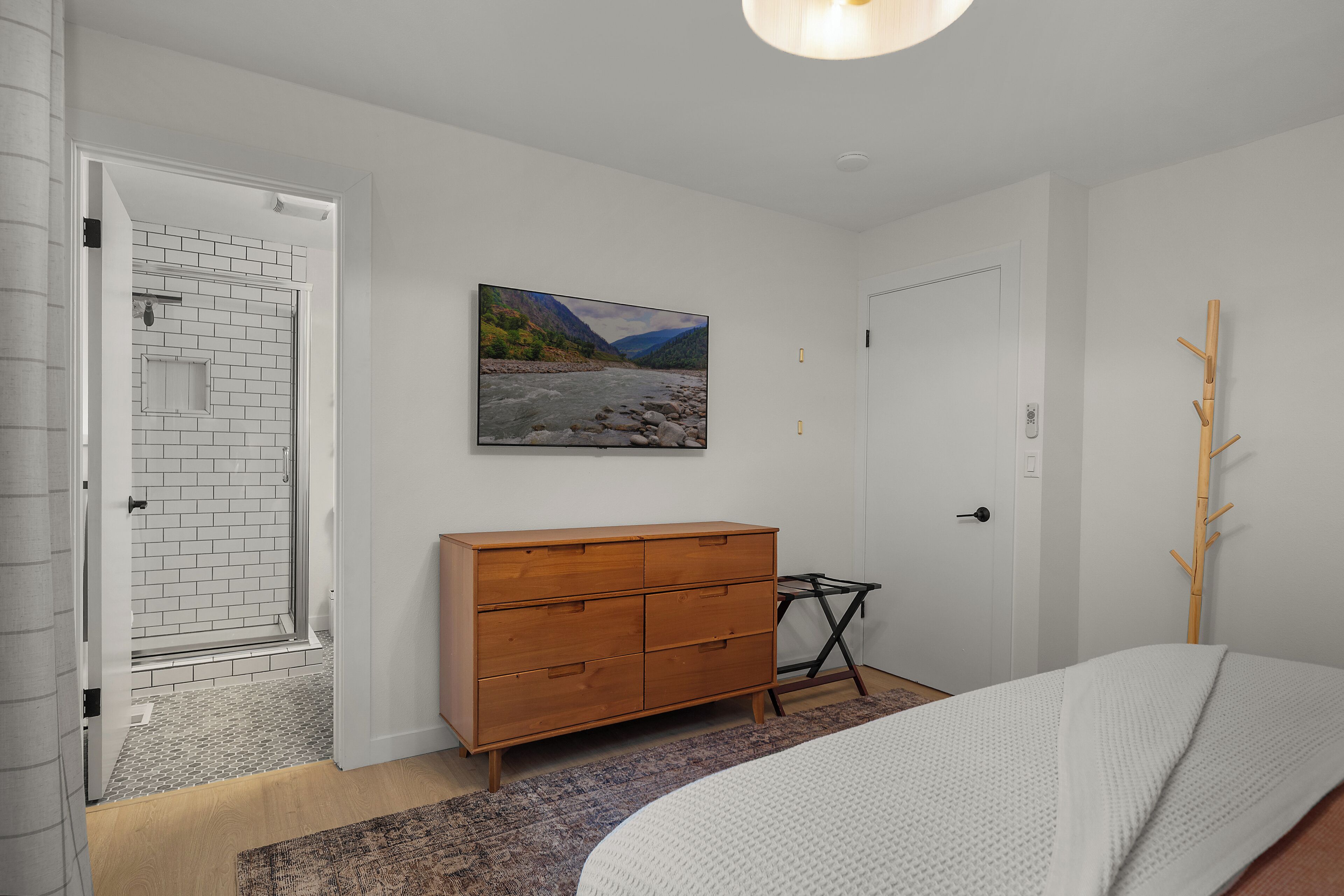 Camera da letto con un letto, una cassettiera in legno, un quadro e una porta che dà su un bagno con doccia in vetro.