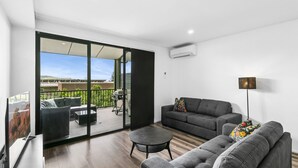 En moderne stue med sofa, sofabord og fjernsyn. Der er en balkon med grill og udsigt over byen.
