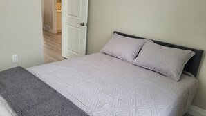 A bed with a patterned bedspread and pillows, a gray throw blanket, and a door leading to a kitchen area. - Small Town Charm (Clifford)