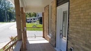 A wooden porch with a metal railing, a brick wall, and a white door. - Small Town Charm (Clifford)