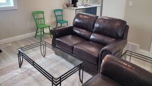 A living room with a brown leather sofa, a glass coffee table, and two green chairs. - Small Town Charm (Clifford)