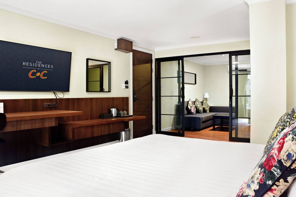A hotel room with a bed, a TV, a wooden desk, a mirror, and a sliding glass door leading to a seating area.