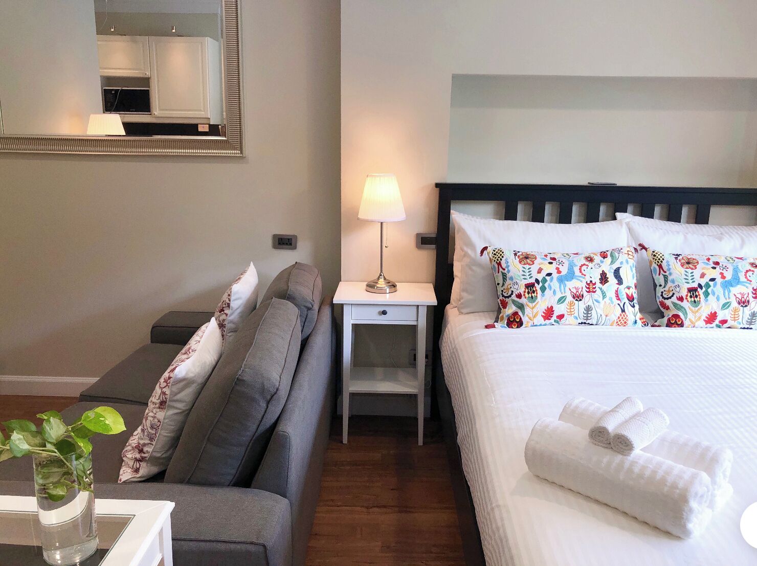 A hotel room with a bed, a sofa, a nightstand with a lamp, and a mirror on the wall.