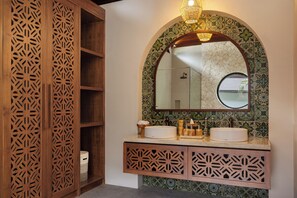 Une salle de bain avec un meuble en bois, un miroir rond et un lavabo agrémenté d’un pare-éclaboussures décoratif en carreaux.