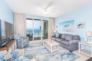 A room with a sofa, a coffee table, a TV, a ceiling fan, and a view of the ocean through a sliding door. - Majestic Sun — Gulf Views, Heated Pool + Hot Tubs (Miramar Beach)