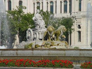 Une fontaine classique ornée d’une statue de femme et de lions, entourée de verdure, avec un bâtiment à colonnes en arrière-plan.