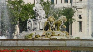 Una fuente clásica con una estatua de una mujer y leones, rodeada de follaje y un edificio con columnas al fondo.