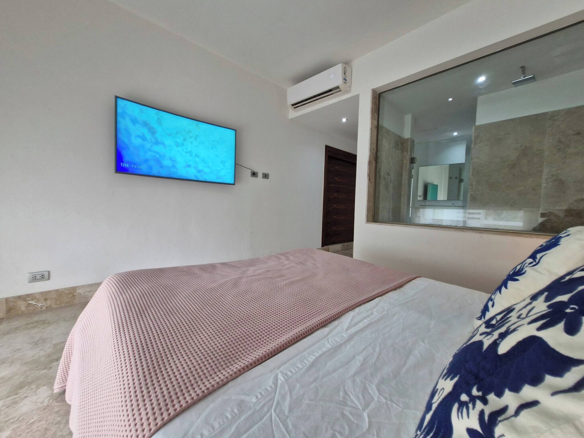 A bedroom with a bed, a TV mounted on the wall, an air conditioning unit, and a large window.