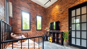 Una habitación con pared de ladrillo, una cama con estructura metálica, un televisor sobre un soporte y un ventanal grande.