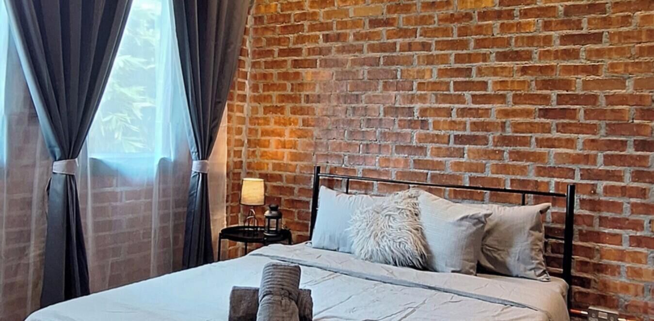 A room with a brick wall, a bed with pillows, a nightstand with a lamp, and a window with curtains.
