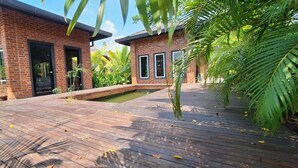 Un edificio de ladrillo de dos pisos con un deck de madera, un pequeño estanque y plantas tropicales.