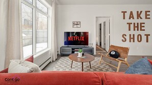 Un salón con una televisión mostrando 'NETFLIX', un sofá, un sillón, una mesita y una pared con el texto 'TAKE THE SHOT'.