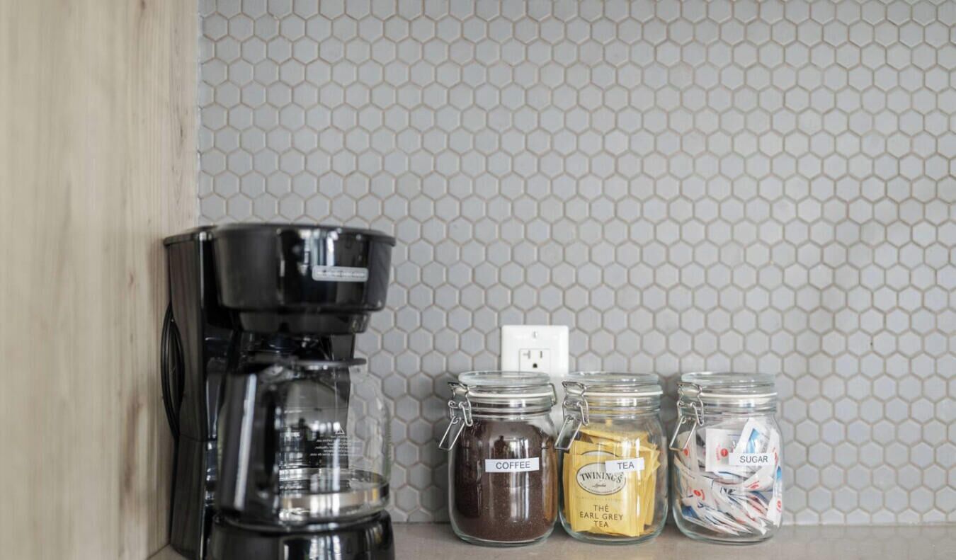 A kitchen with a coffee maker, two glass jars labeled coffee, tea, and sugar, and a hexagonal tiled backsplash.