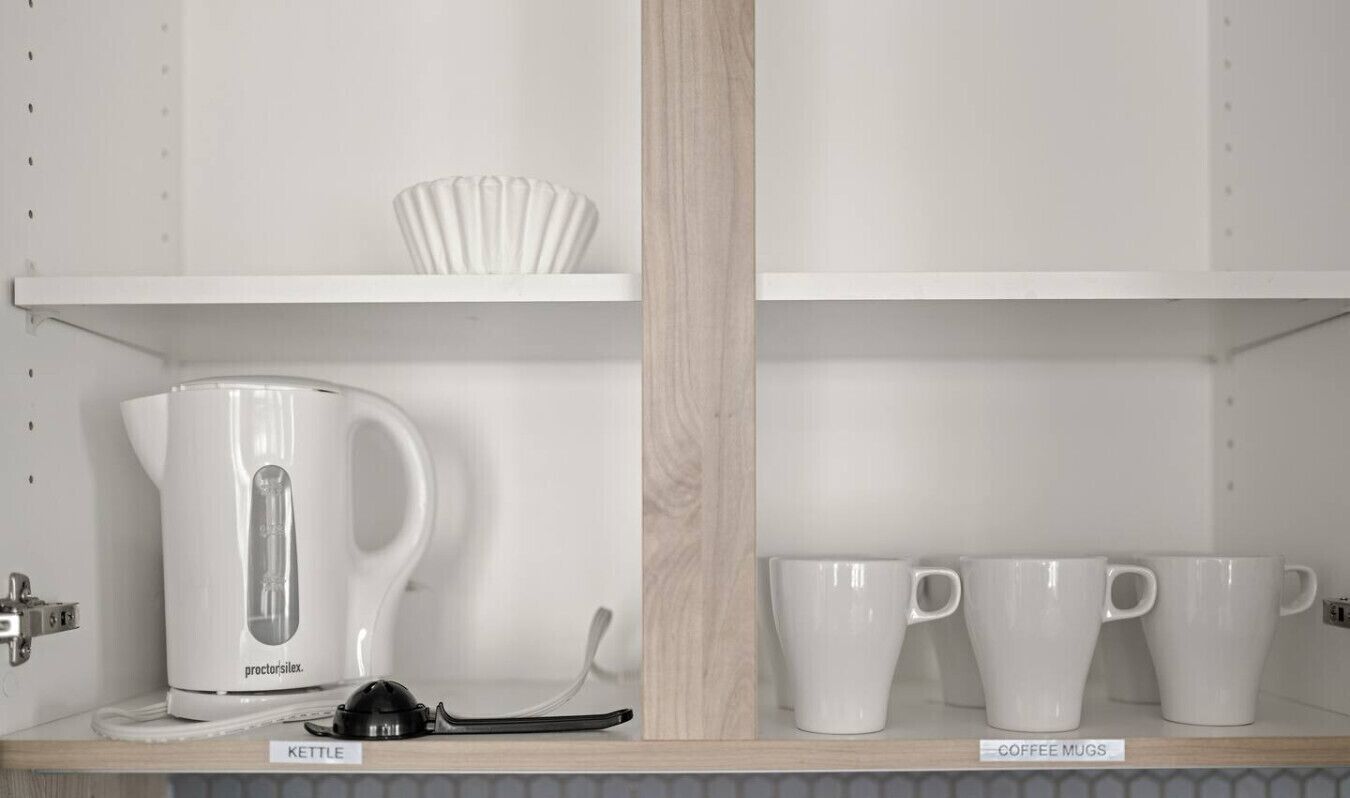A kitchen shelf with a kettle, coffee mugs, and a spoon.