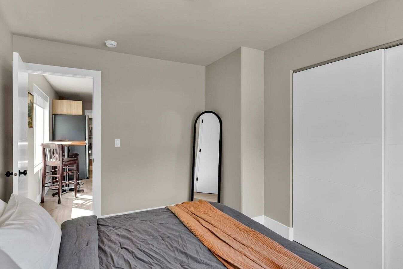 A bedroom with a bed, a large mirror, and a wardrobe. There is a door leading to a kitchen area.