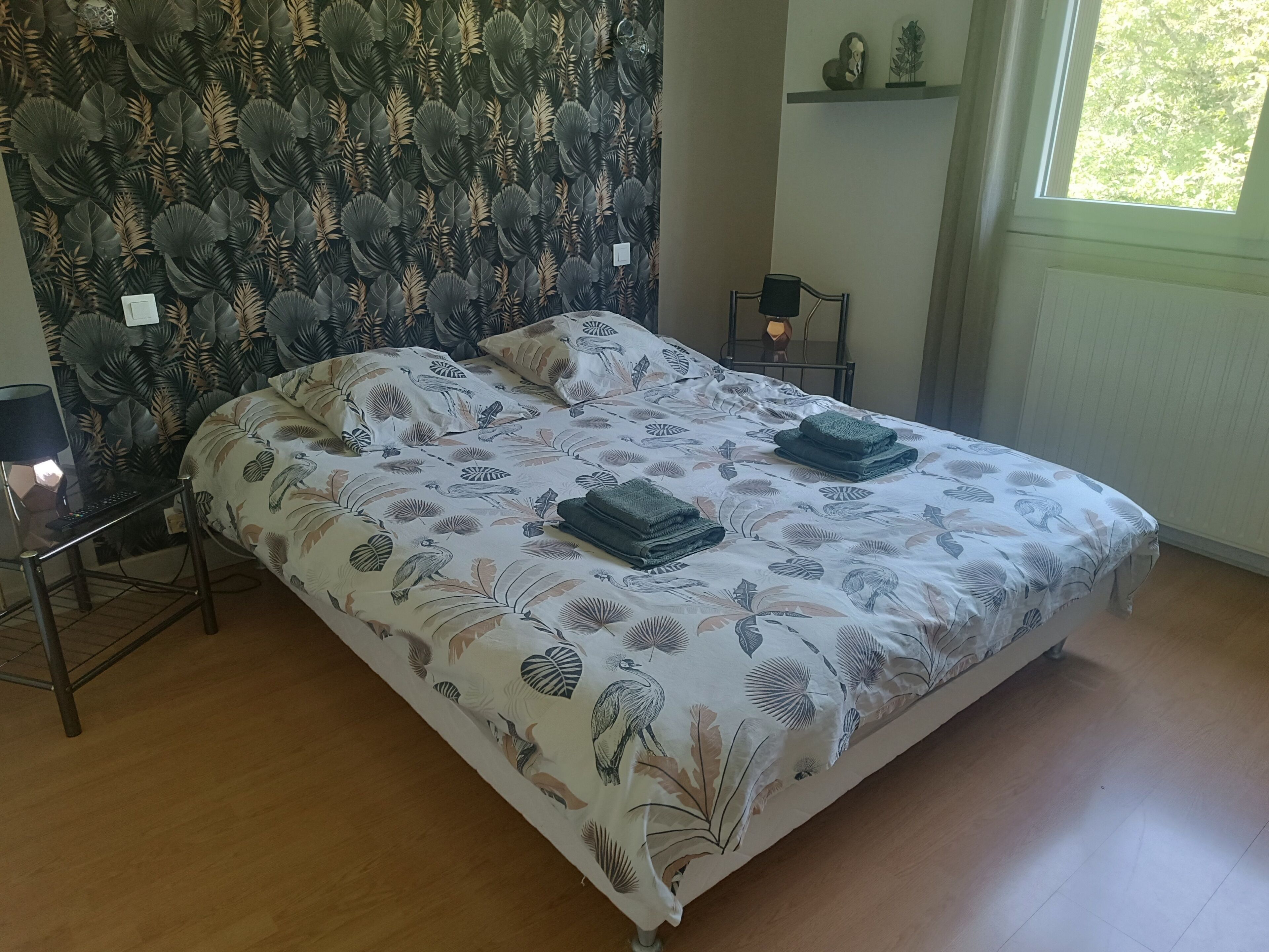 A bedroom with a bed, a bedside table, a lamp, a wall with a leaf pattern, and a window.