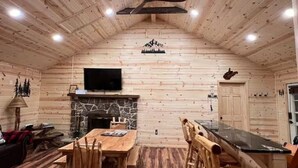 Ein Interieur einer Blockhütte mit Kamin aus Naturstein, einem Holzespeisesaal und einem an der Wand befestigten Fernseher.