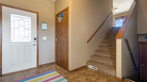 Un pasillo con una puerta blanca, una puerta de madera, una escalera con barandilla y una alfombra colorida.