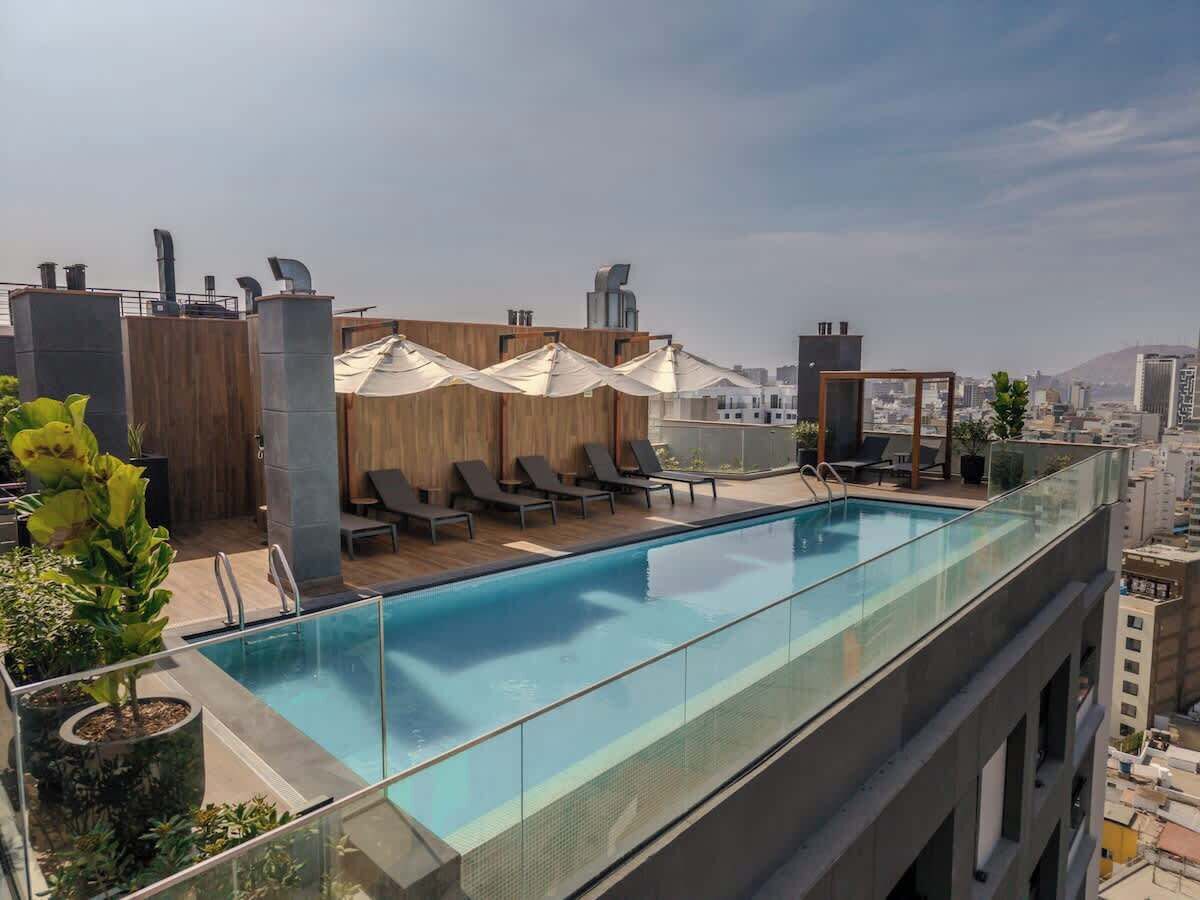 A rooftop pool with lounge chairs and umbrellas, surrounded by greenery and city views.