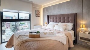 Una camera da letto con una grande finestra, una testiera trapuntata, un comodino con una lampada e un letto con coperta e cuscini color beige.