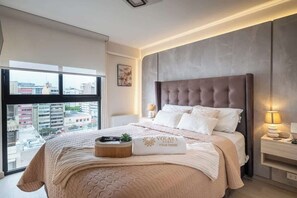 A bedroom with a large window, a tufted headboard, a bedside table with a lamp, and a bed with a beige blanket and pillows. - Modern and cozy apartment in Larco Miraflores. (Miraflores)