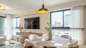 A modern living room with a large flat-screen TV, a dining area with a round table, and a view of the city through large windows. - Largest & Elegant Apartment | Larco | Miraflores (Miraflores)