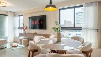 A modern living room with a large flat-screen TV, a dining area with a round table, and a view of the city through large windows.
