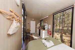 Een slaapkamer met een bed, een houten bankje, een kleine kitchenette en uitzicht op een bos.