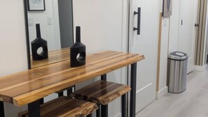 A modern hotel room with a wooden dining table, black metal stools, a mirror, and a trash bin. - The Loft Moden Rental (Calgary)