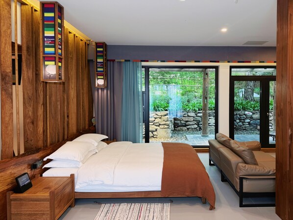 A modern bedroom with a bed, bedside tables, a sofa, and a view of a garden with a waterfall.