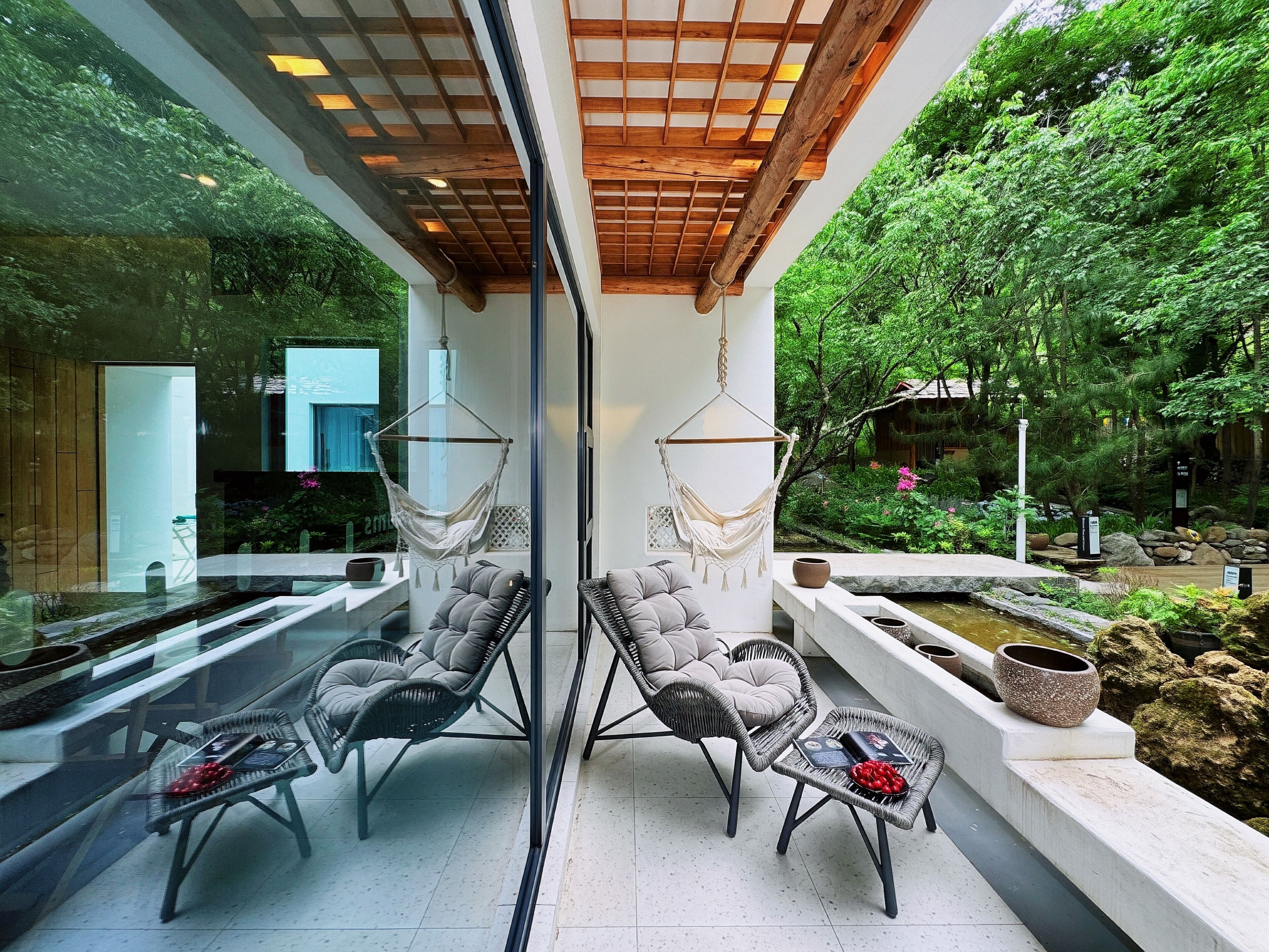 A modern outdoor seating area with cushioned chairs and a hanging chair, overlooking a lush garden and a water feature.