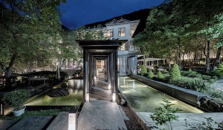 A modern building with a covered walkway, water features, and lush greenery.. Between Trees and Streams
