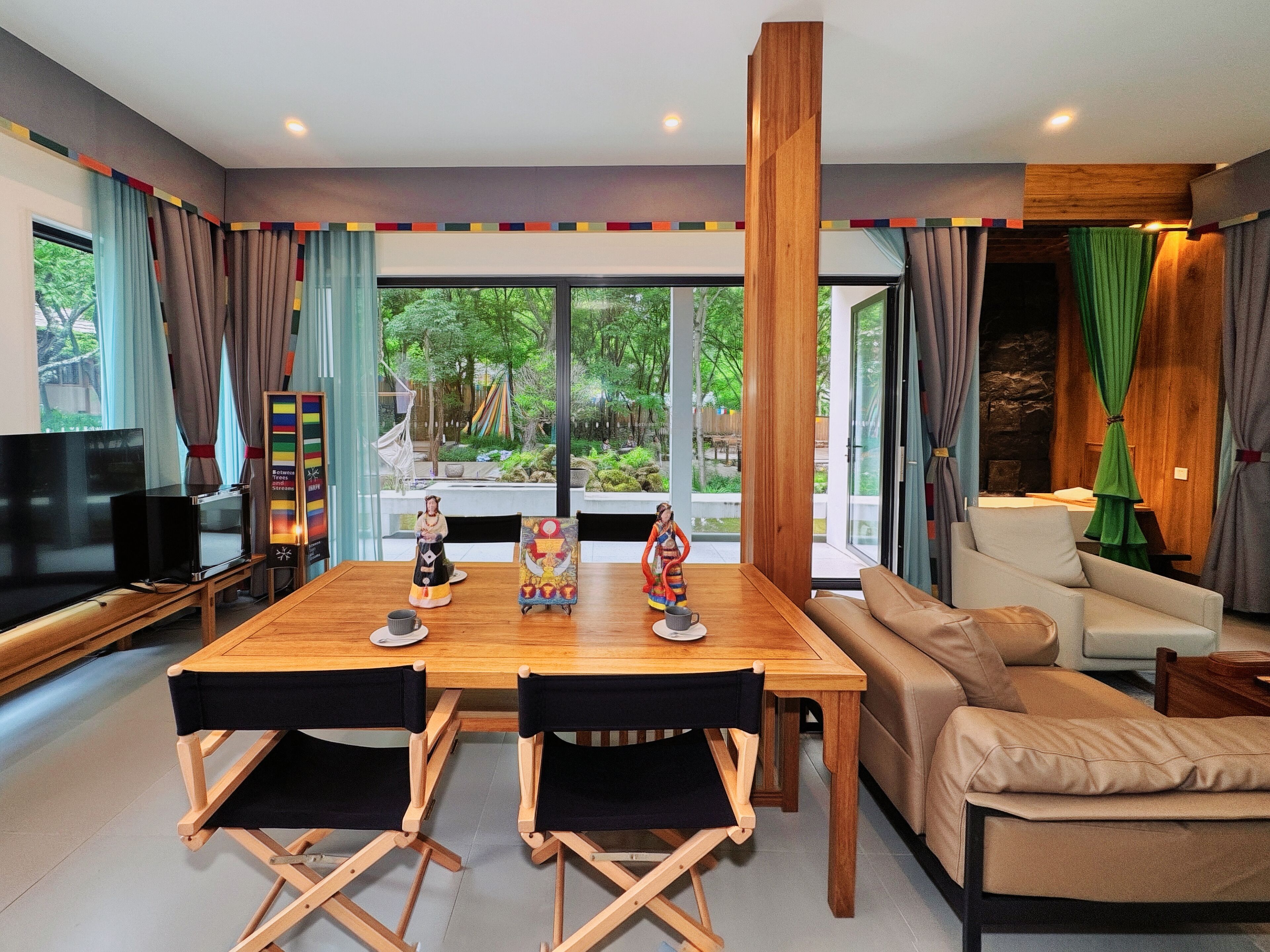 Un salon moderne avec une table à manger en bois, deux chaises noires, un canapé, un téléviseur et une grande fenêtre donnant sur un paysage verdoyant.