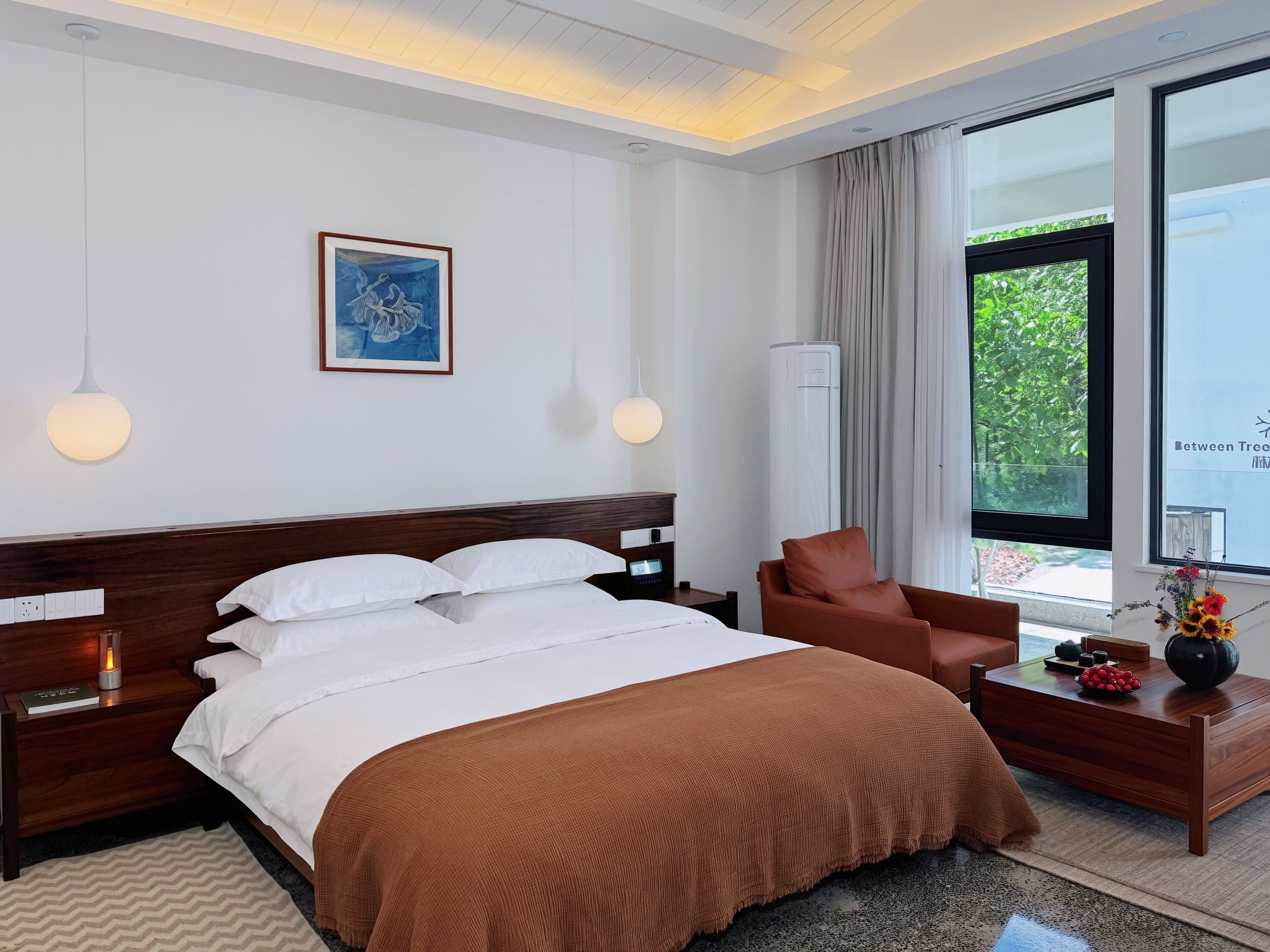 Habitación de hotel con cama, una silla, mesita de noche, lámpara, un cuadro y ventana con vistas.