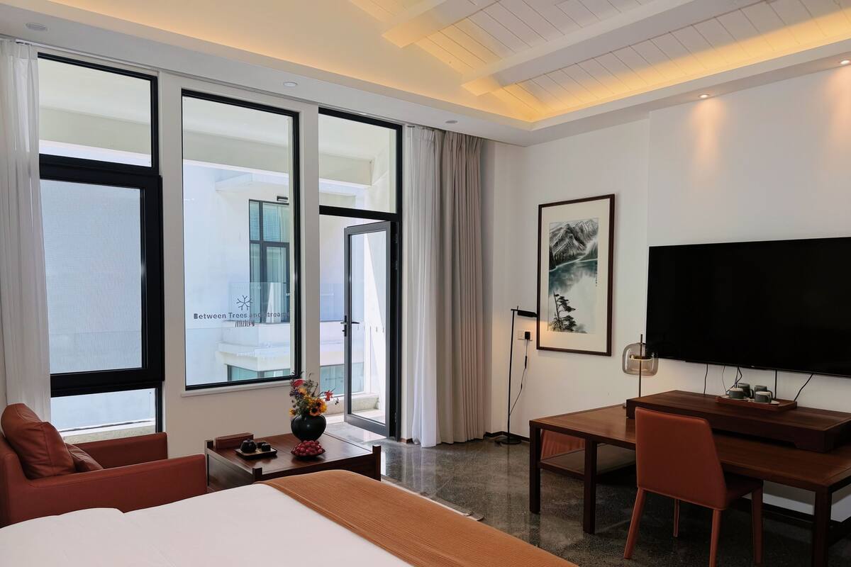 A hotel room with a bed, a desk, a chair, a TV, and large windows.