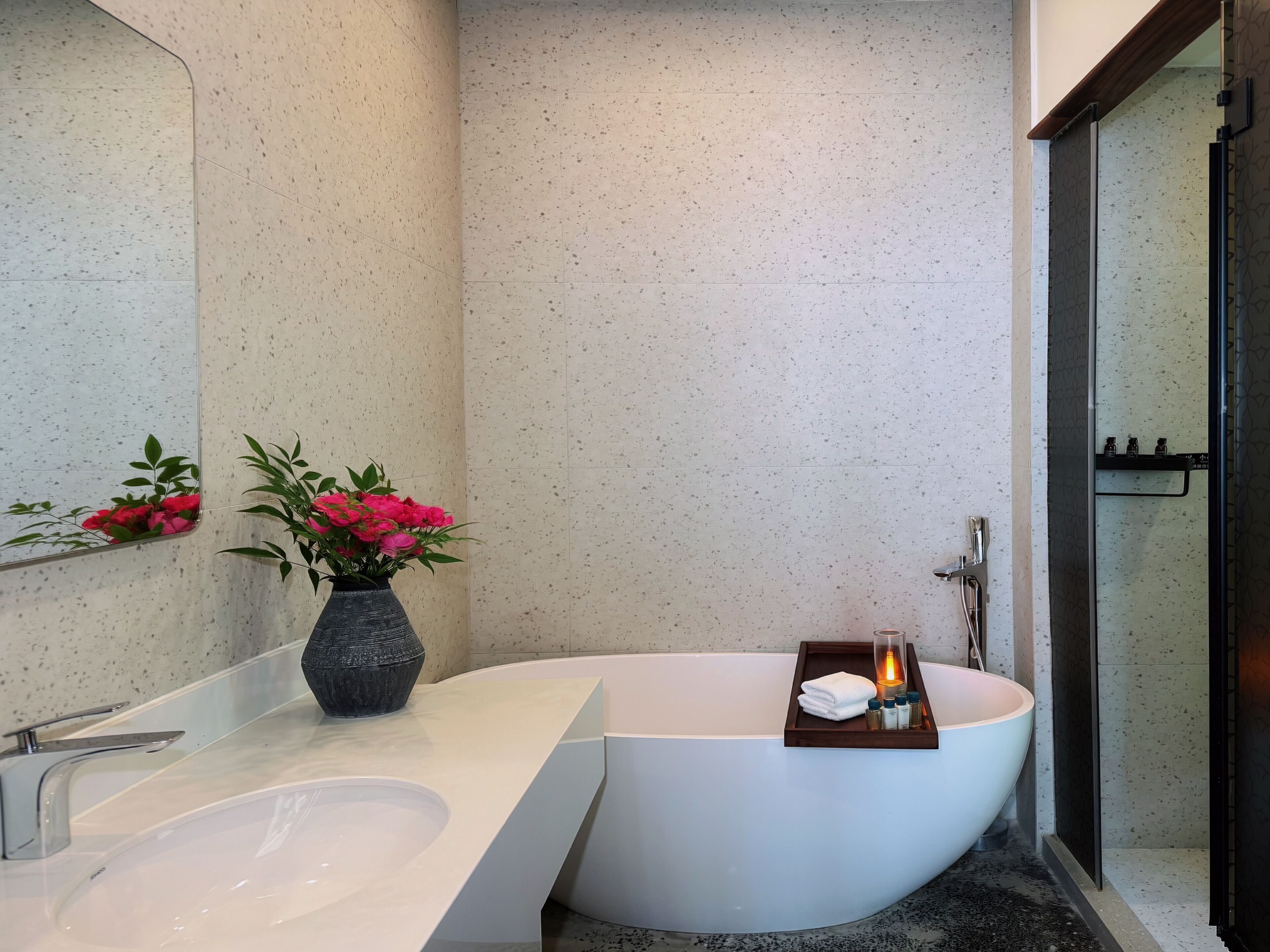 A modern bathroom with a freestanding bathtub, a large mirror, and a small vase with flowers.