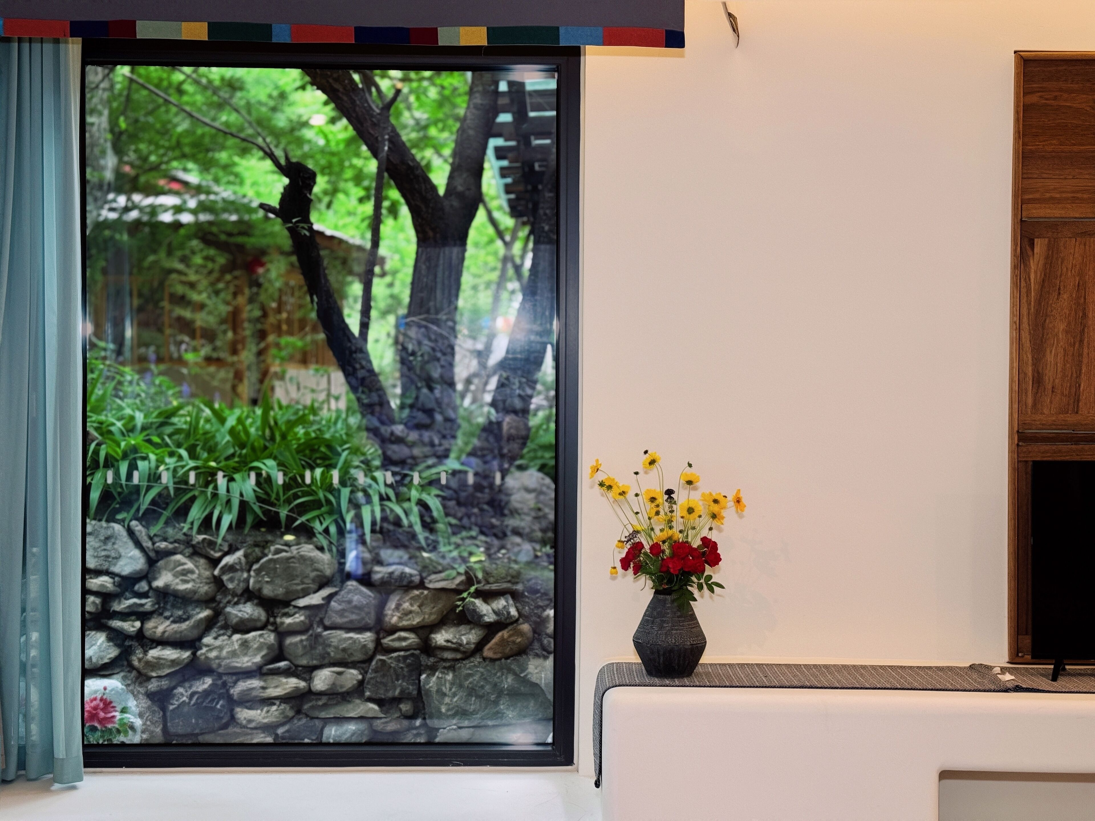 A room with a large window, a stone wall, and a vase with flowers on a table.
