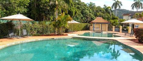 A clear swimming pool surrounded by lush greenery and a few buildings.