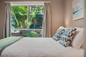 A bedroom with a bed, curtains, a window with a view of greenery, and a painting on the wall. - Splash at Sapphire - Pet Friendly, Resort Pool, Beachfront (Coffs Harbour)