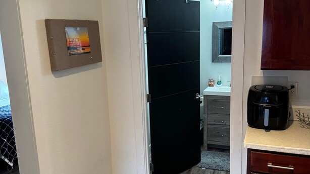 Un couloir avec une porte noire, un tableau encadré au mur, une machine à café sur le comptoir et un plancher en bois.
