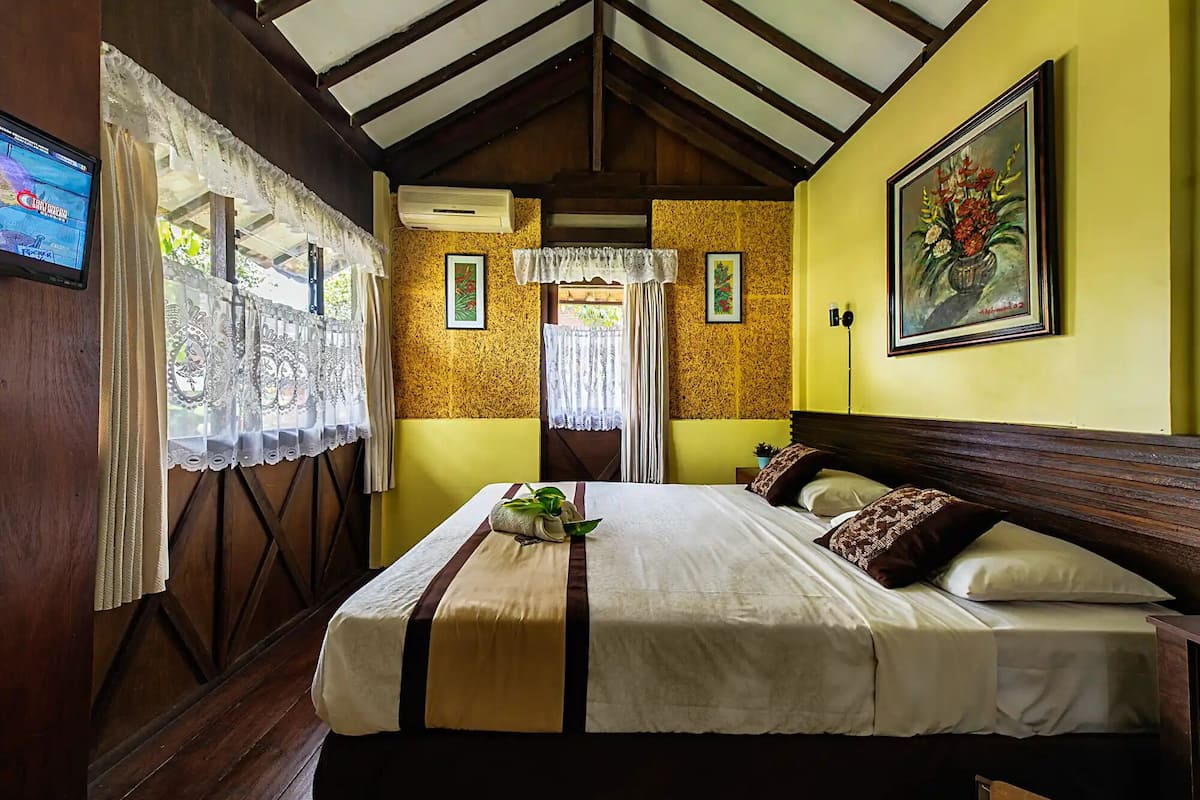A bedroom with a bed, a window with lace curtains, a painting on the wall, and an air conditioning unit.