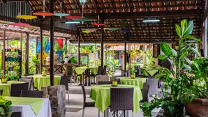 An indoor restaurant with tables covered in green tablecloths, wicker chairs, and hanging colorful umbrellas. - Dedaunan Guesthouse (Batu)